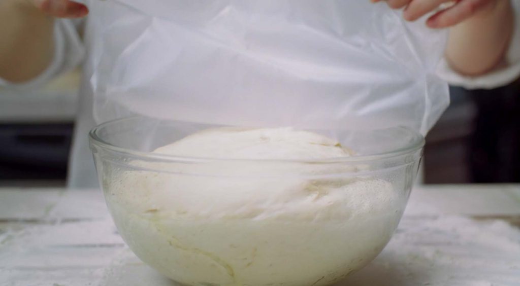 Dough rising in a clear glass bowl, covered with plastic wrap. The dough is ready to be transferred to a proofing basket for further fermentation.